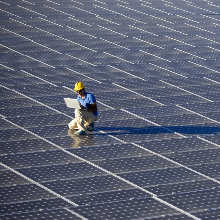 Worker in a yellow hard hat kneeling on a solar panel array, using a laptop to assess operational data and manage potential risks associated with solar farms.