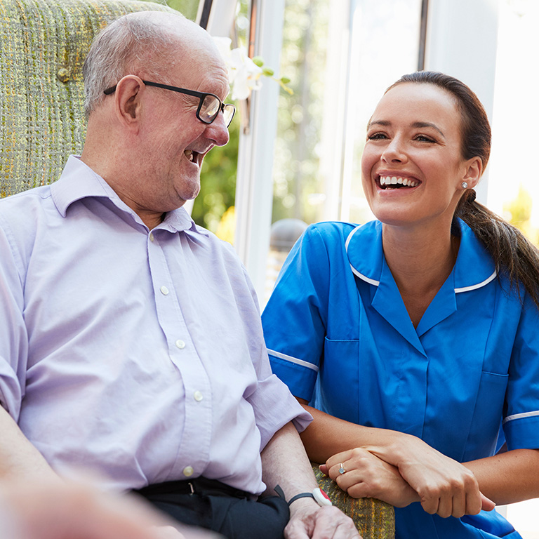 Senior Man Sitting In Chair And Talking With Nurse In Retirement Home