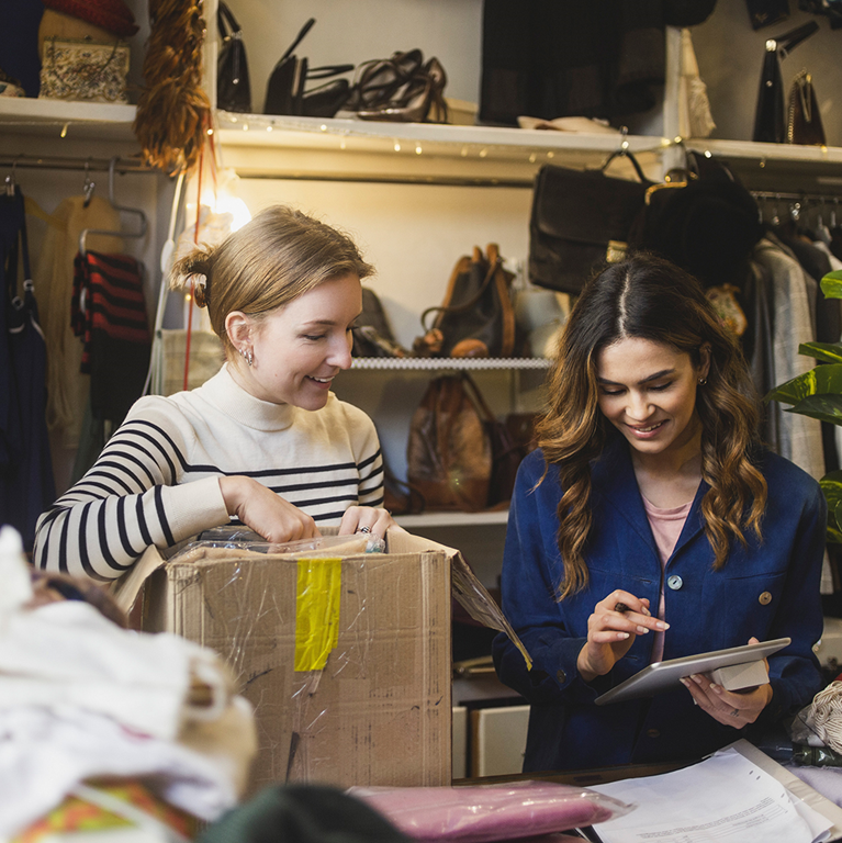 Two women in a retail setting, one examining a package while the other uses a tablet, surrounded by various clothing items and accessories.