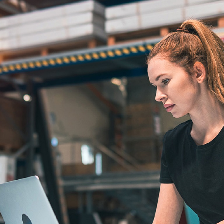 Woman in a warehouse with stacked shelves behind her working on a laptop