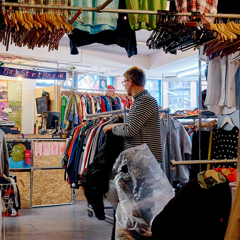 A shop owner sorting clothes on racks in a vibrant thrift store, surrounded by various garments and a welcoming atmosphere.