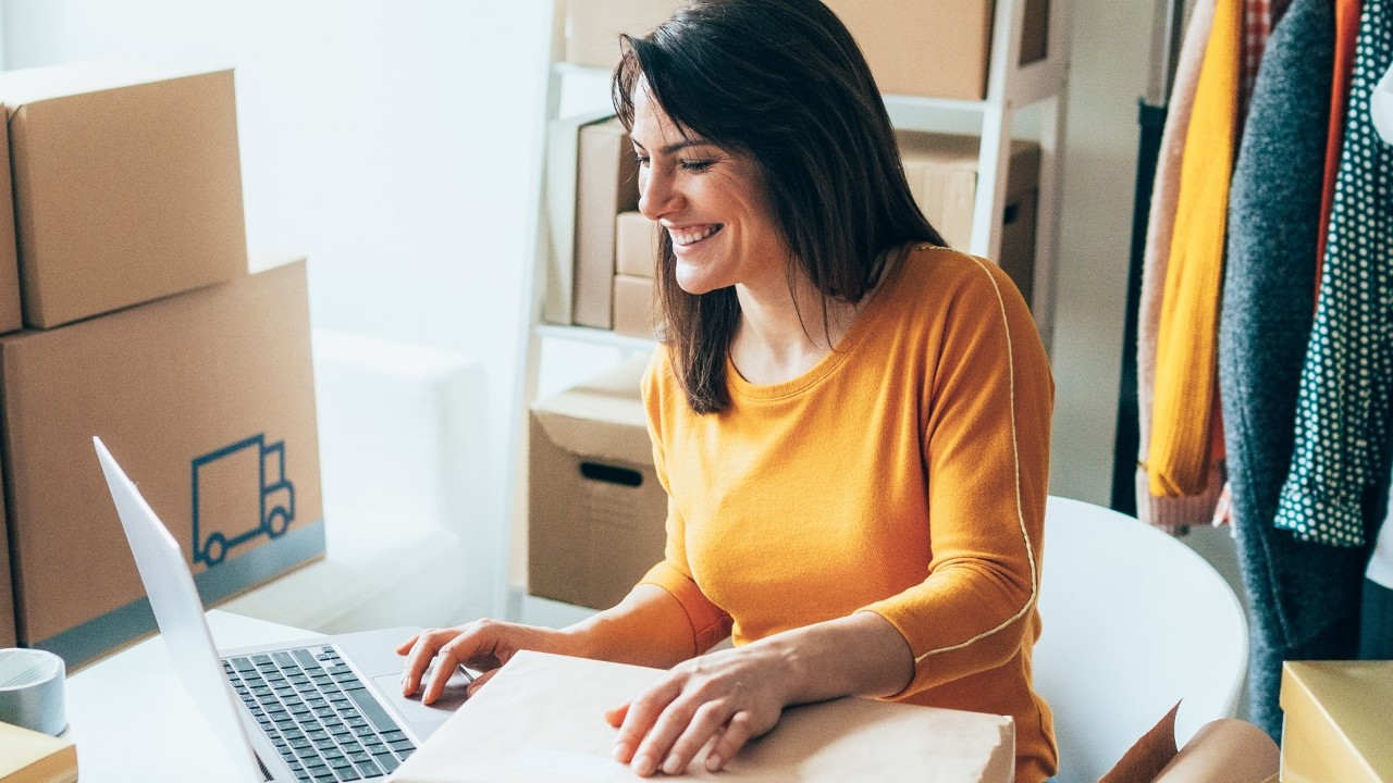 Online retailer smiling, working on a laptop with carboard boxes and clothes rail behind: online retailers insurance